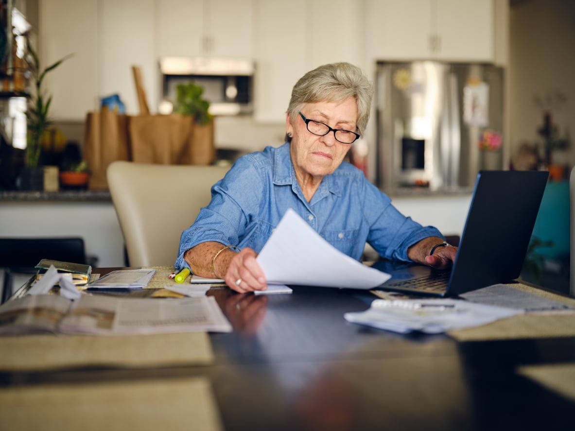 Older woman wearing glasses works at a table with a laptop, holding papers and surrounded by documents in a home kitchen setting, reflecting strong Baby Boomer beliefs in dedication and hard work.