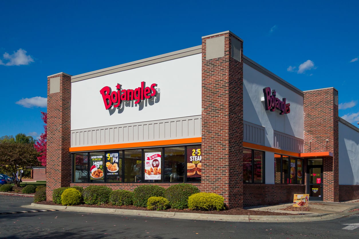 A Bojangles restaurant with a brick exterior, large windows displaying food posters, and a clear blue sky in the background. As one of the popular regional fast-food chains, this location features neatly trimmed shrubs and a spacious parking lot visible in front of the building.