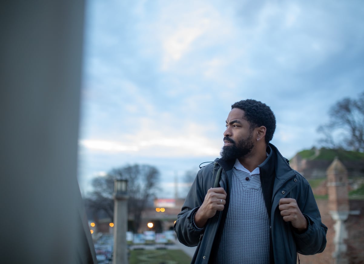 A man with a beard wearing a jacket and backpack stands outdoors at dusk, looking to the side with a thoughtful expression. Blurred city lights and trees appear in the background. Tourist mistakes.