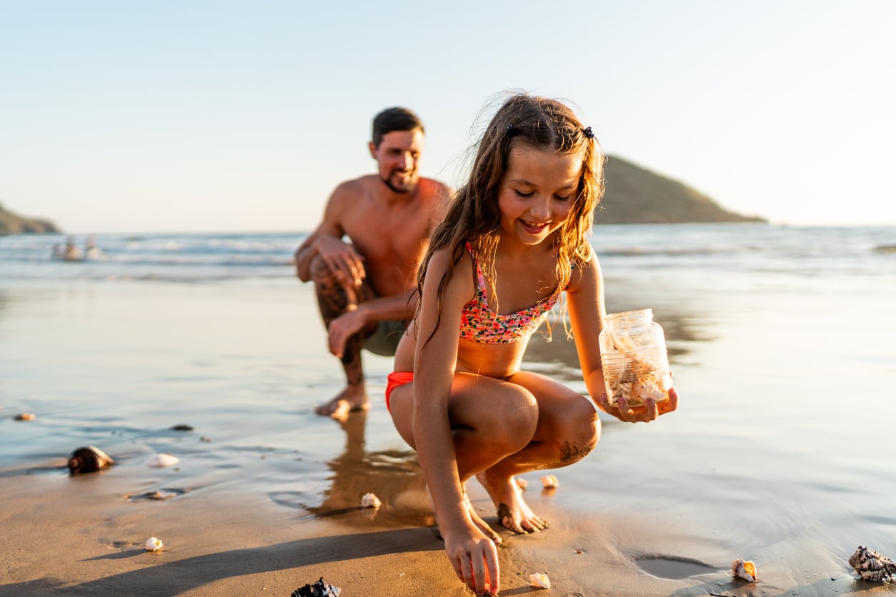 A young girl in a swimsuit collects seashells on a beach while holding a jar, perhaps for her beach vacation savings, as an adult man squats behind her with the sea and hills in the background under a clear sky.