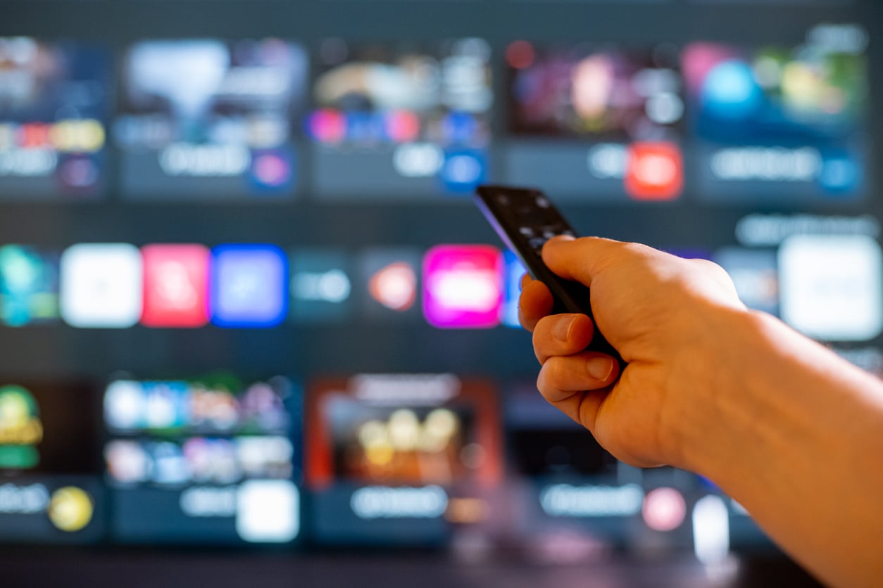 A hand holds a remote control, pointing it at a blurred TV screen displaying various streaming service app icons and colorful content thumbnails, suggesting the practice of money saving habits by carefully choosing which subscriptions to keep.