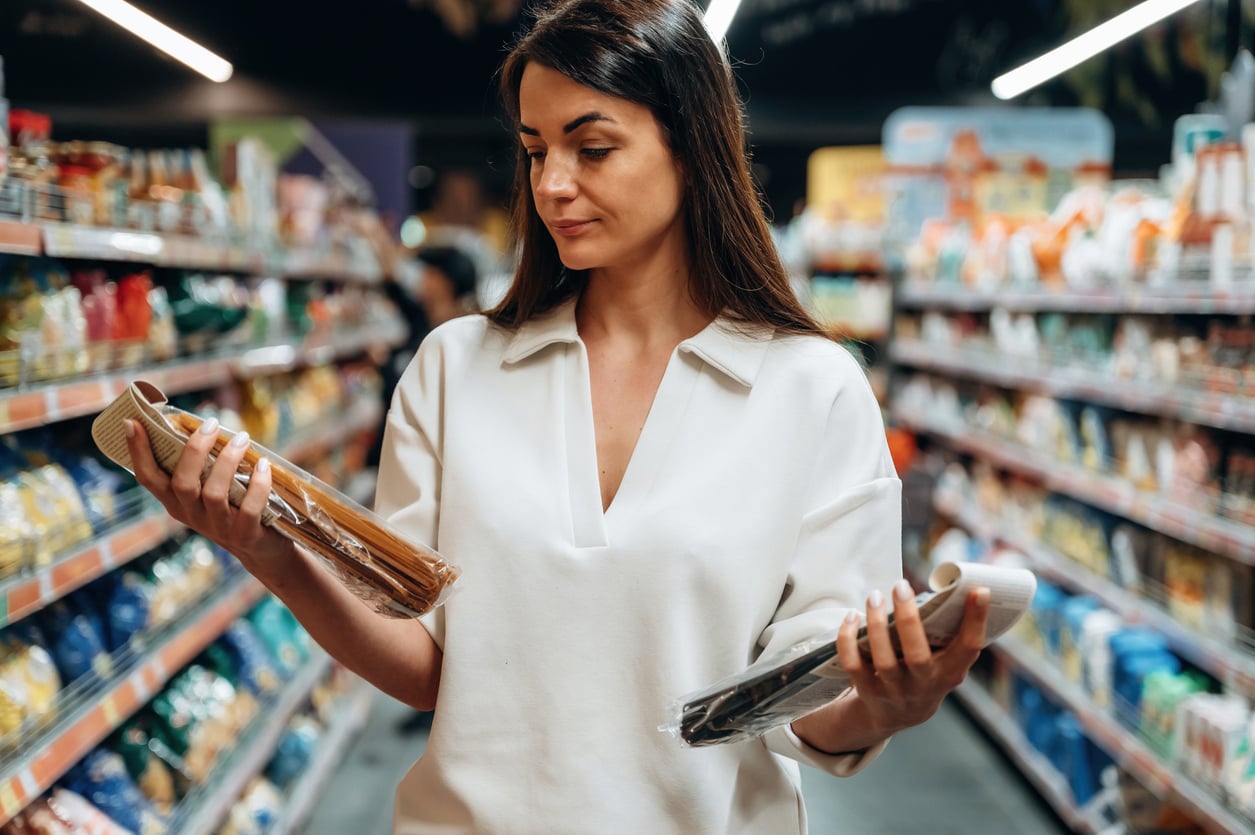 A woman in a white shirt stands in a grocery store aisle, holding and comparing two packaged items while shopping. Shelves stocked with various products are visible in the background, highlighting how women are changing their spending behaviors through thoughtful purchasing decisions.
