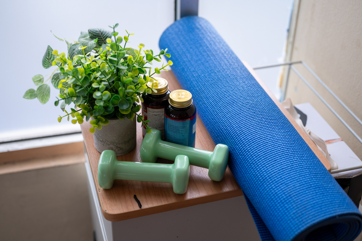 A small table holds a potted green plant, two green dumbbells, two brown pill bottles, and a rolled-up blue yoga mat near a window—perfect for staying healthy at home and save money on gym memberships.