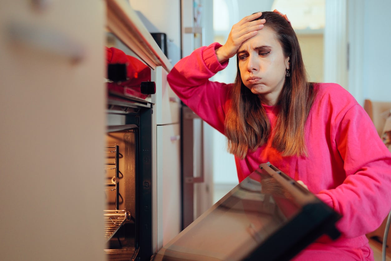 A woman in a pink sweater sits in front of an open oven, holding her forehead in frustration, exhaling deeply—her expression reveals disappointment or stress, perhaps worried about household expenses tied to her cooking or baking.