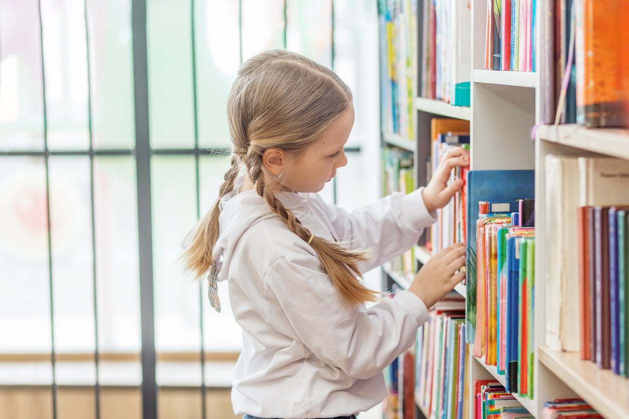 A young girl with braided hair and a white shirt selects a book from a shelf in a brightly lit library or bookstore.