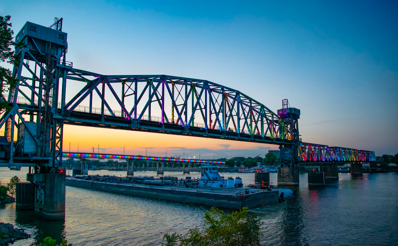 A cargo barge passes under a large steel lift bridge with colorful lights at dusk, reflecting on the river below—a tranquil scene often admired in picturesque beach towns. The sky is a gradient of blue and orange.