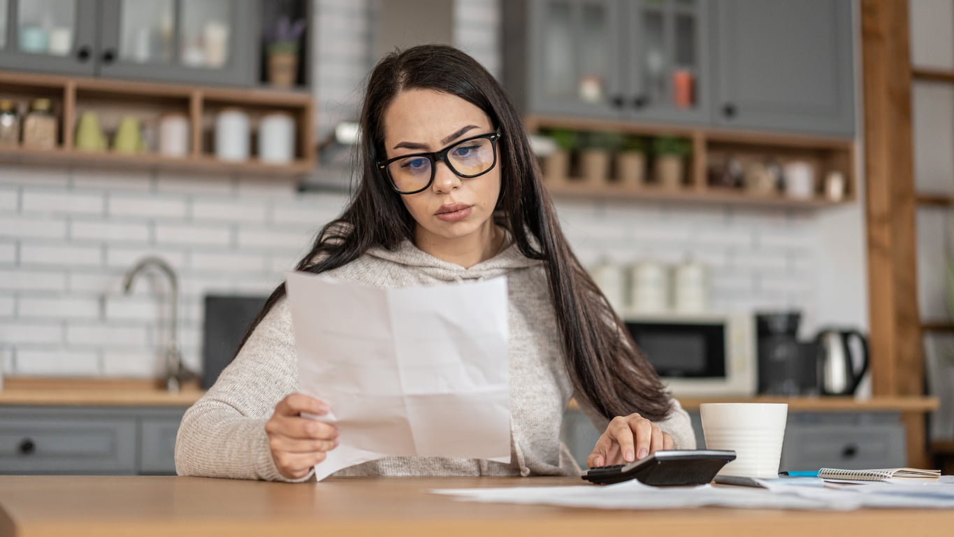 A woman with long dark hair and glasses sits at a kitchen table, looking at a piece of paper. She appears focused and concerned as she reviews household expenses, with a calculator, papers, and a coffee cup in front of her.