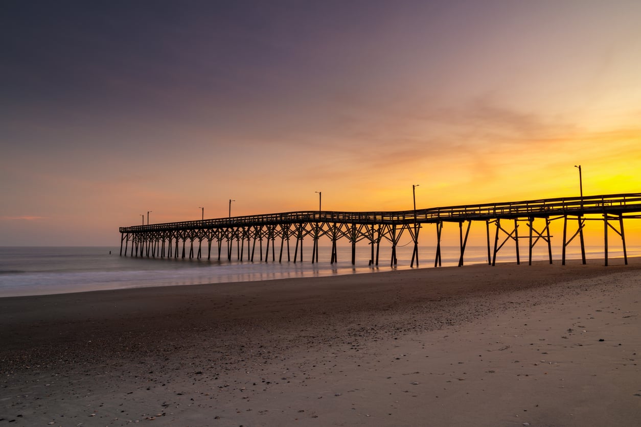 A long wooden pier stretches over calm water at sunrise with a glowing orange and purple sky. The sandy beach in the foreground, reminiscent of classic beach towns, has scattered shells and gentle waves.