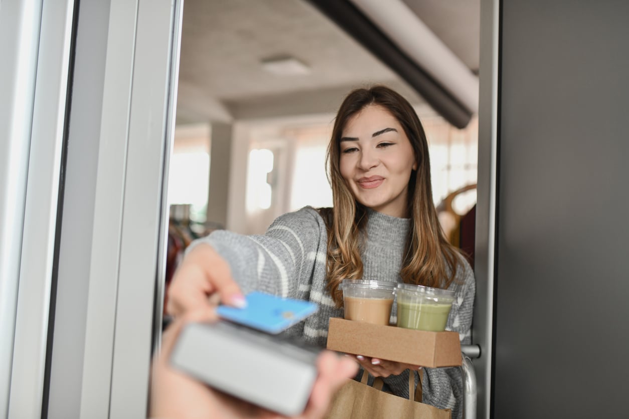 A woman at a door smiles while handing a credit card to someone. She is holding cups of drinks and a paper bag, suggesting she is receiving a delivery.