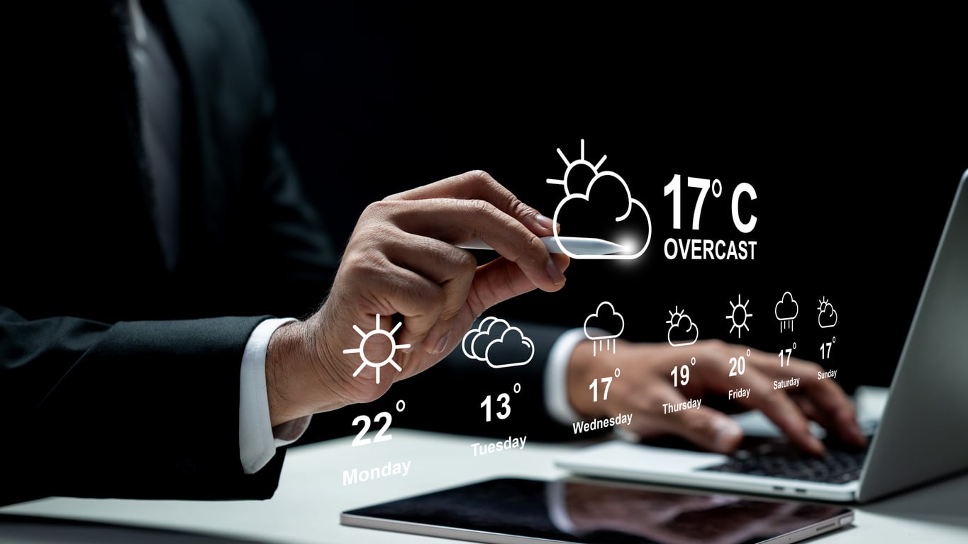 A person in a suit uses a laptop with virtual weather icons projected, showing a weekly forecast with temperatures and conditions—including overcast at 17°C—while preparing a road trip checklist.