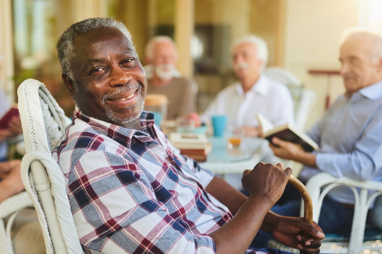 Smiling older man in a plaid shirt sits in a wicker chair holding a cane, embodying classic Baby Boomer beliefs about friendship and community, with three other elderly men talking and reading books in the blurred background.