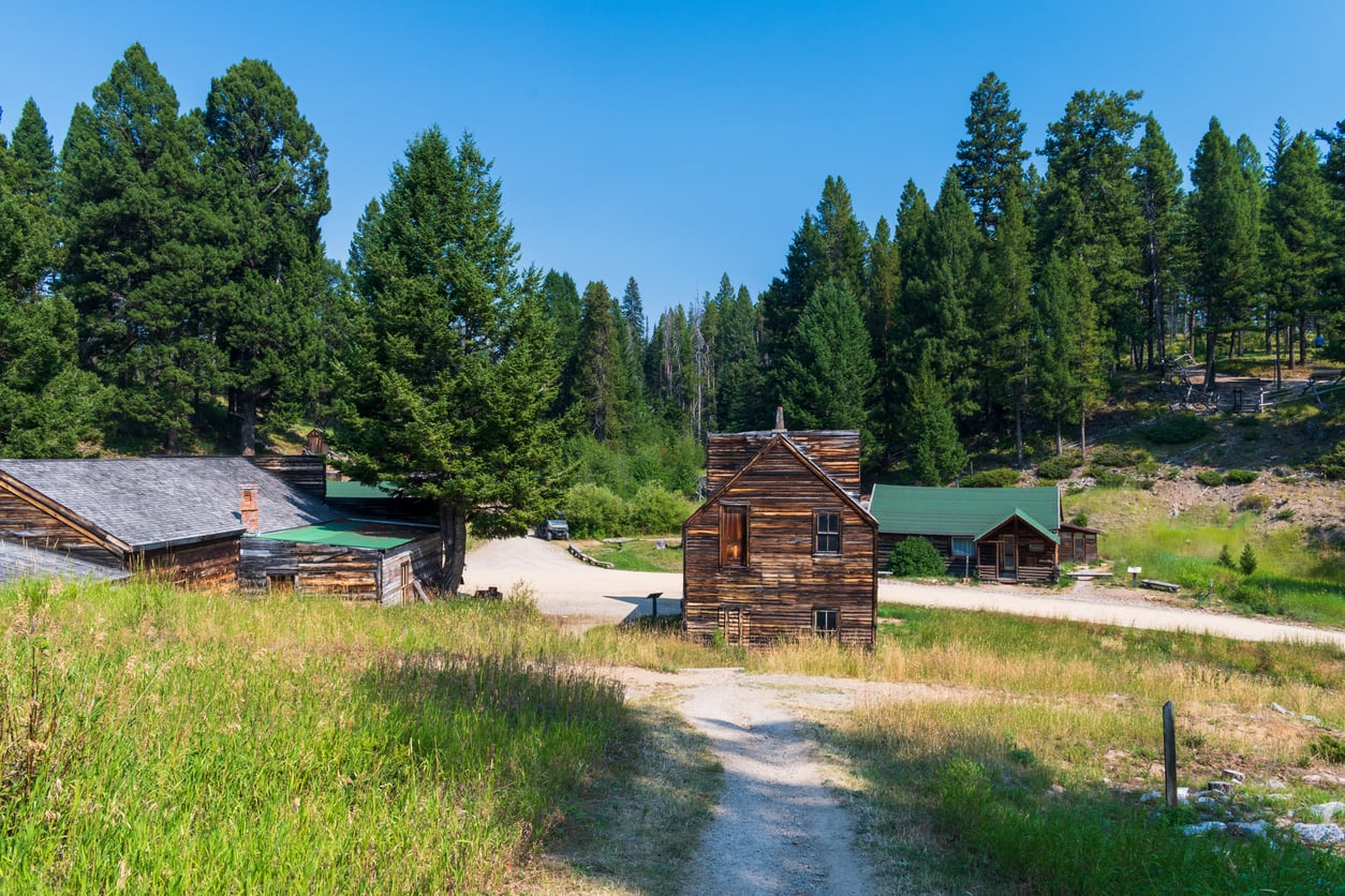 A dirt path leads to rustic wooden cabins surrounded by trees and grassy hills under a clear blue sky, evoking the timeless charm of american towns nestled in a rural, forested area.