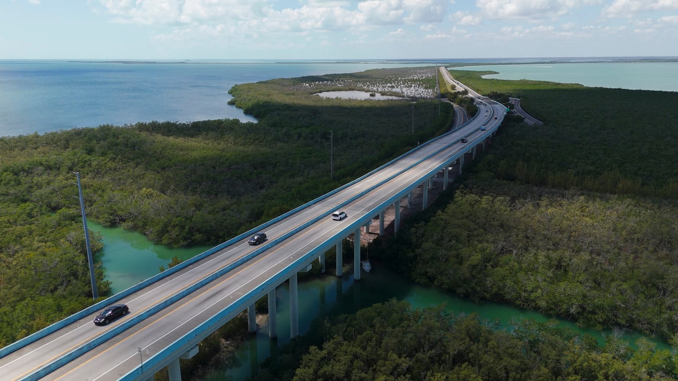 Aerial view of cars driving on a long bridge over turquoise water, surrounded by green trees and wetlands, with a coastline and blue sky in the background.