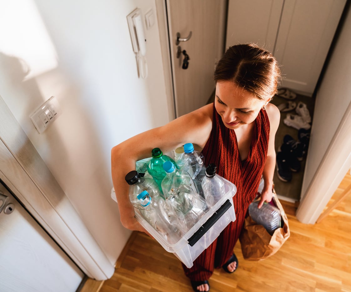 A woman in a red dress, perhaps mindful of vacation costs, carries a clear bin filled with empty plastic bottles and a paper bag as she leaves a room with wooden floors and shoes by the door.