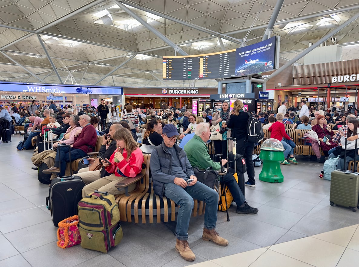 A busy airport departure lounge with many people sitting and waiting, surrounded by luggage. Shops and a Burger King are visible in the background, and a large departures board hangs above.