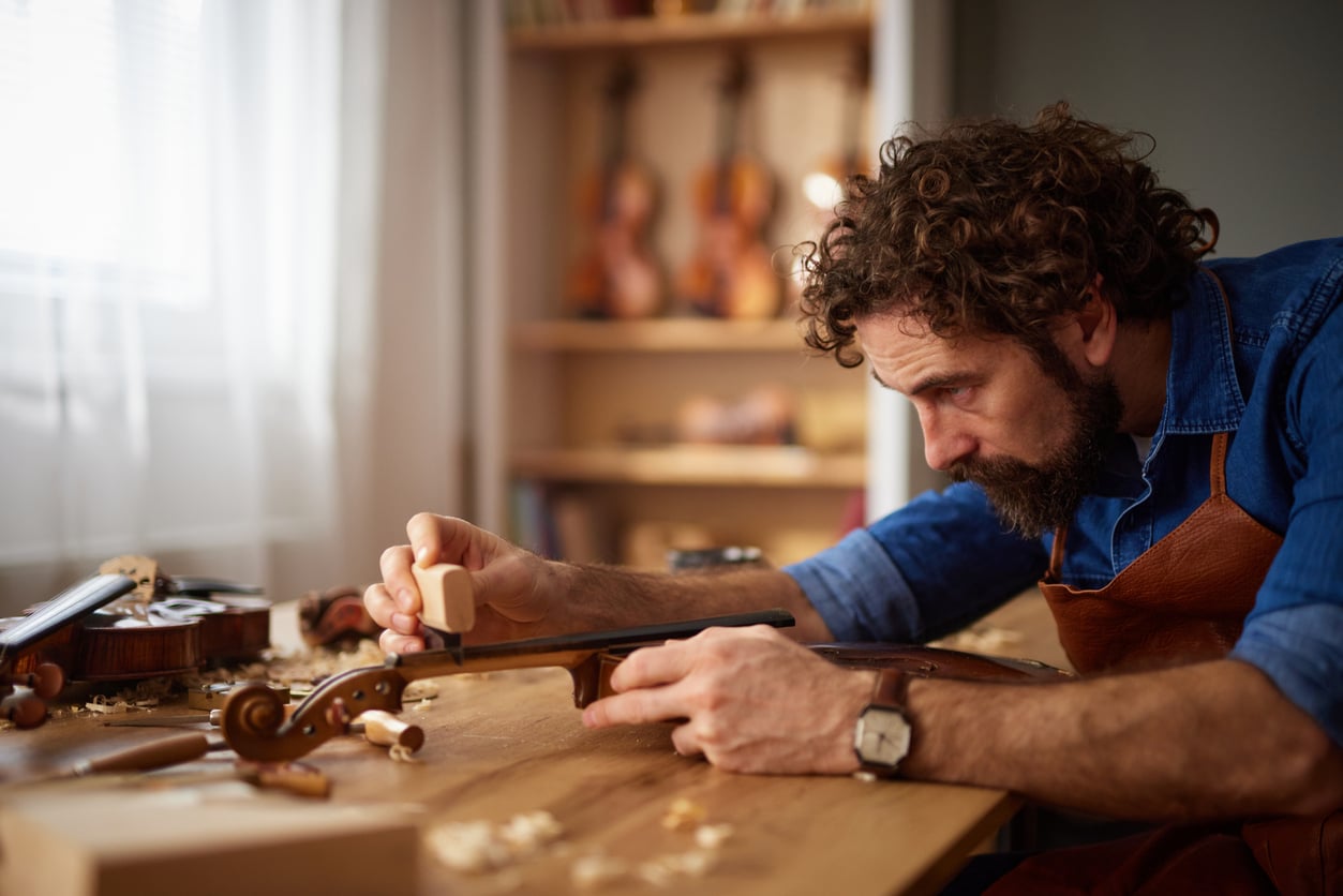 A man with curly hair and a beard carefully repairs a violin at a wooden workbench in a workshop filled with tools and other string instruments, reflecting the meticulous dedication often associated with expensive hobbies.