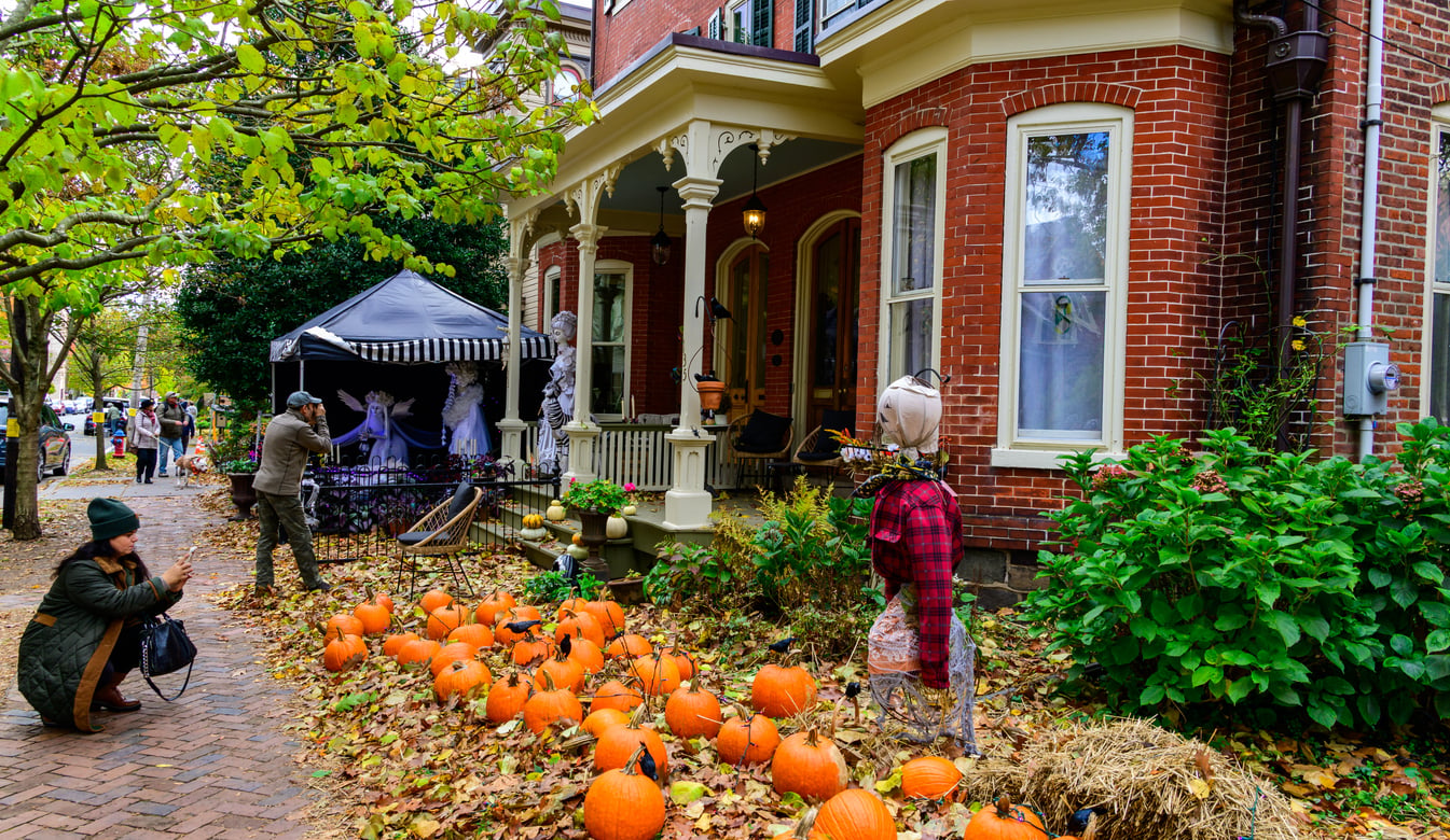 A woman kneels to photograph pumpkins and fall decorations outside a brick house with a porch, while others walk nearby. Scarecrows and autumn leaves enhance the festive outdoor scene in one of the best trick or treat towns in America.