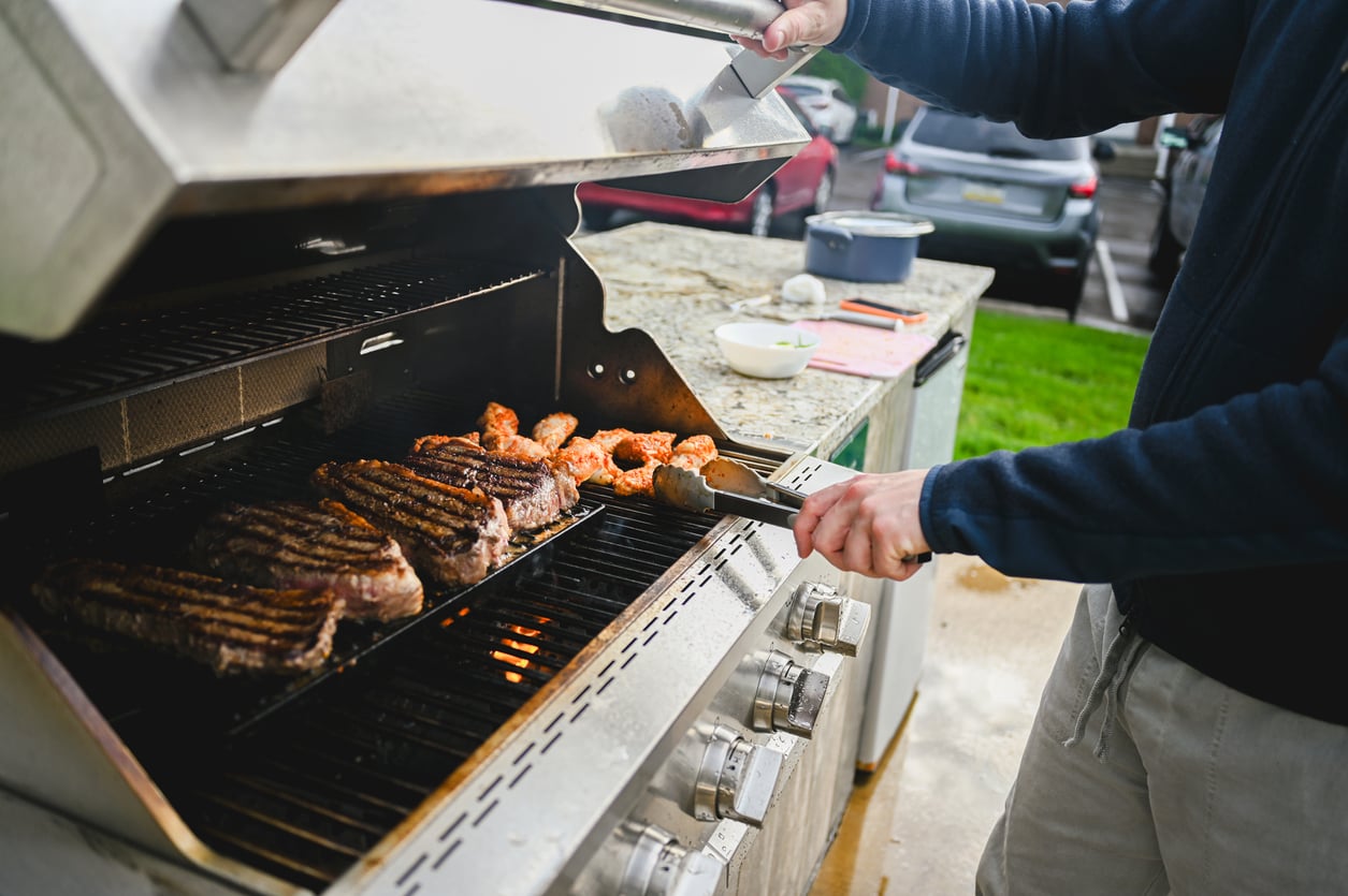 A person grills steaks and chicken on a large outdoor gas barbecue, showcasing one of the most popular outdoor home products. The grill lid is open, and the person uses tongs to turn the meat while cars and grass are visible in the background.