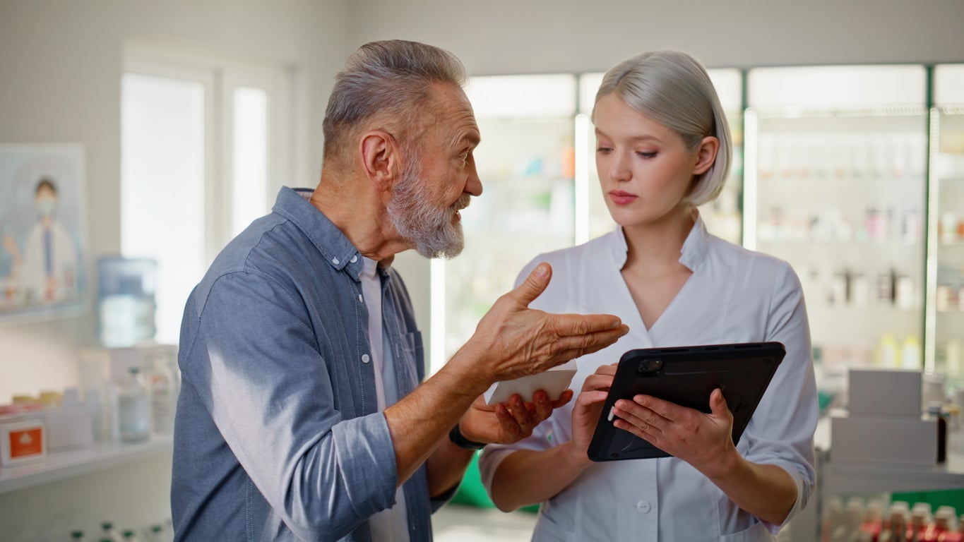 An older man, embodying classic Baby Boomer beliefs, gestures while speaking to a young pharmacist holding a tablet in a pharmacy. Both appear engaged in conversation, surrounded by shelves with various medicines.