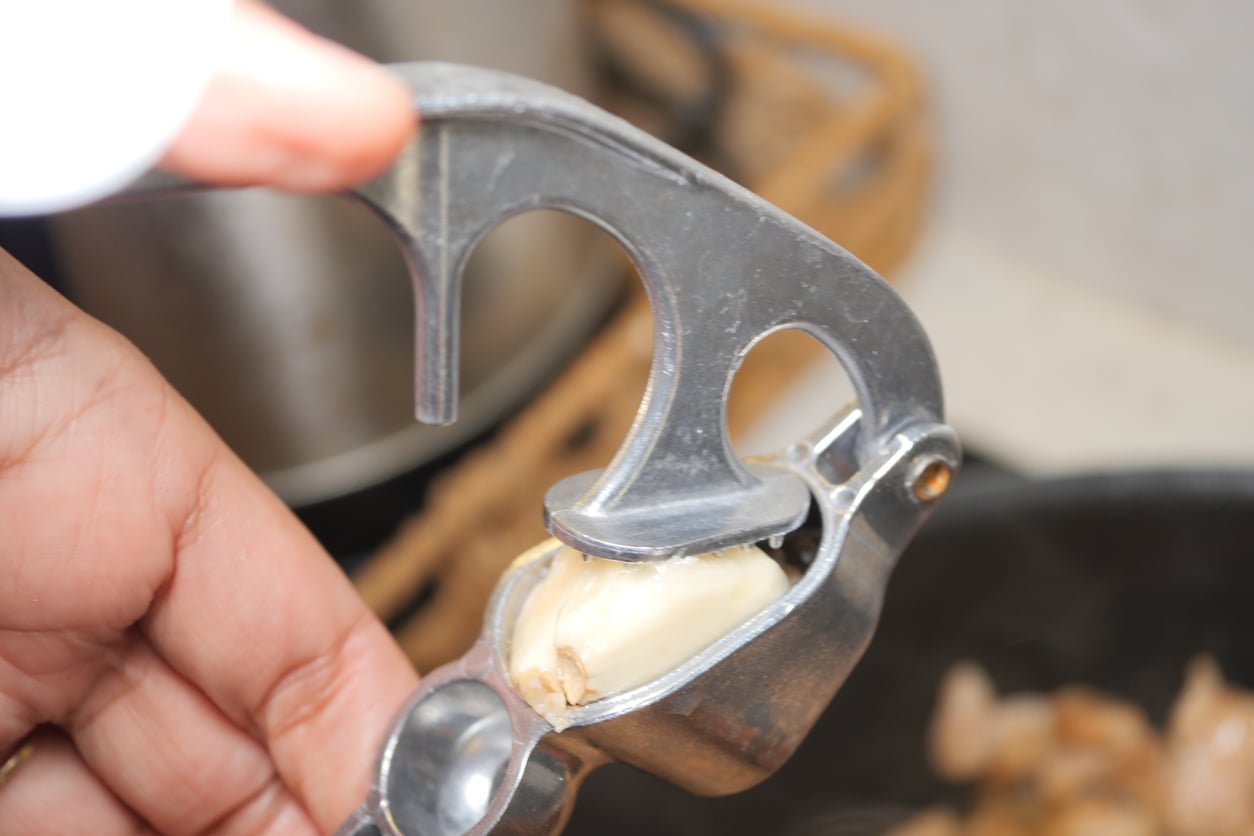 A hand holds a metal garlic press with a peeled garlic clove inside, ready to be crushed over a pan in a kitchen setting—though some might argue that garlic presses are among the more common waste of money products found in home kitchens.