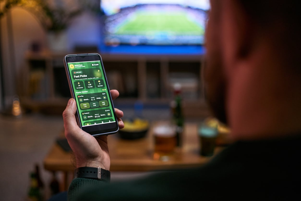 A person holds a smartphone displaying a sports betting app while watching a game on TV, with drinks and snacks on a table in the background, demonstrating how money saving habits can play a role even while enjoying entertainment.