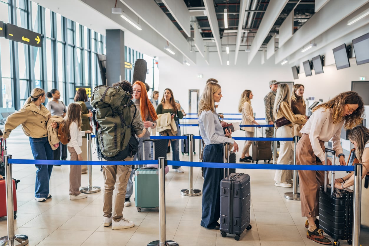 Travelers stand in line at an airport check-in area, with luggage beside them. Some people are checking documents or opening suitcases, while others wait. The terminal is bright with large windows and overhead monitors.