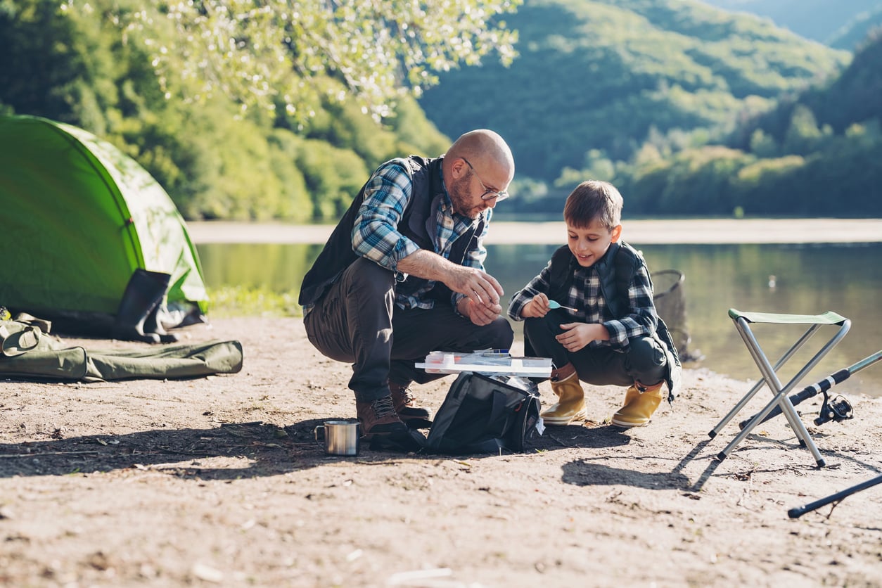 A man and a boy sit by a lake, preparing fishing gear near a green tent with mountains and trees in the background. The boy smiles as the man helps him, enjoying the outdoor setting and sharing one of their favorite—if sometimes expensive—hobbies together.