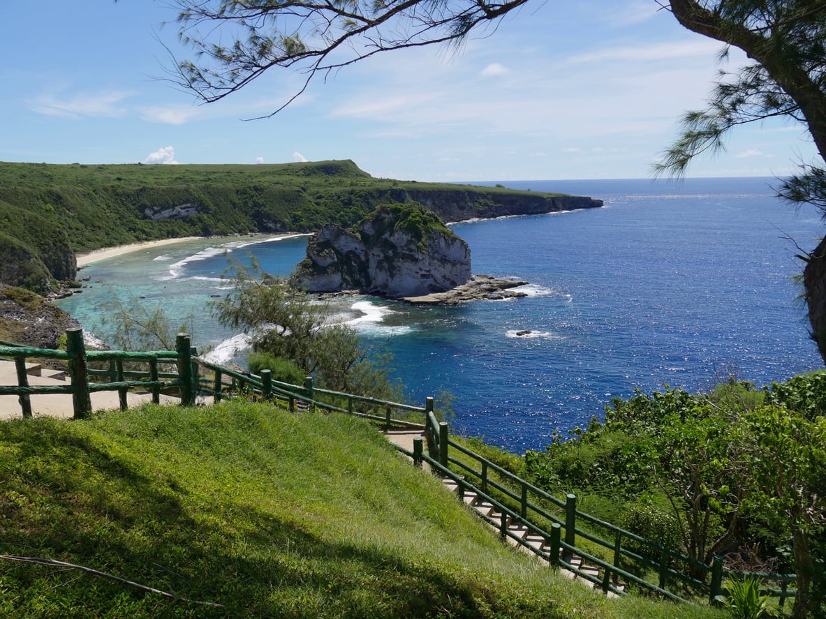 A scenic view of a blue ocean beside a rocky coastline, green cliffs, and a winding path with a wooden fence, framed by lush trees under a clear sky—one of the breathtaking travel destinations perfect for nature lovers and explorers alike.