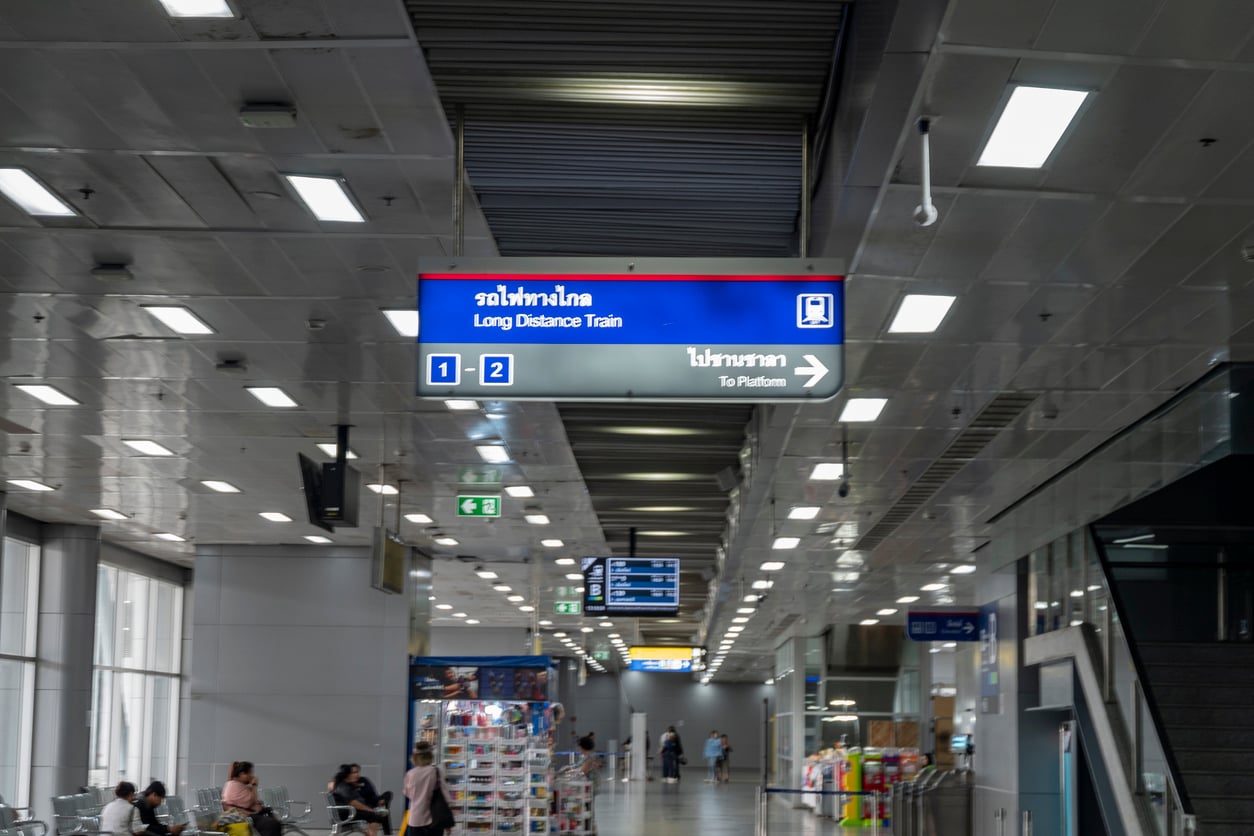 A blue and white sign in a train station directs passengers to platforms 1 and 2 for long distance trains. People are sitting and walking in the bright, modern hall, perhaps considering their travel options before booking a flight.