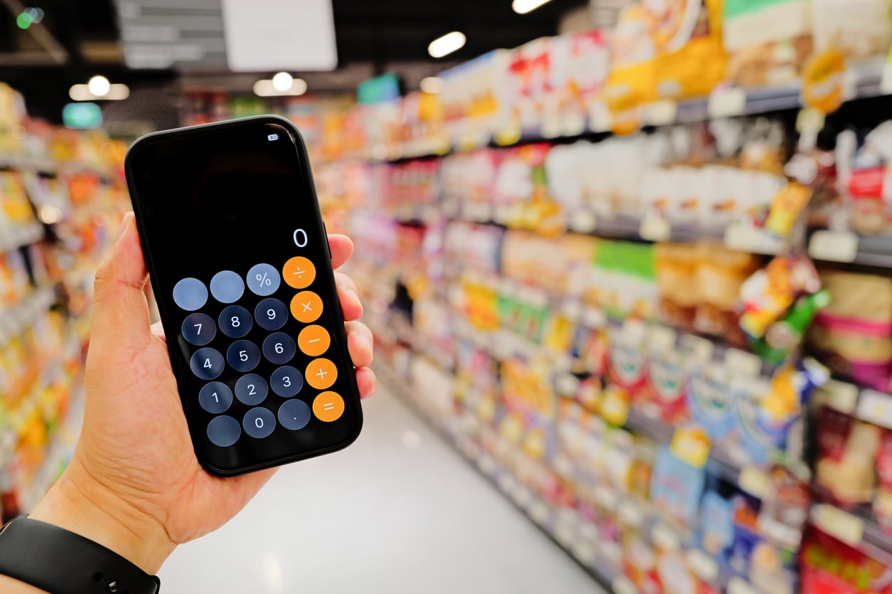 A hand holds a smartphone with a calculator app open, displaying zero, in front of blurred shelves stocked with groceries in a brightly lit supermarket aisle.