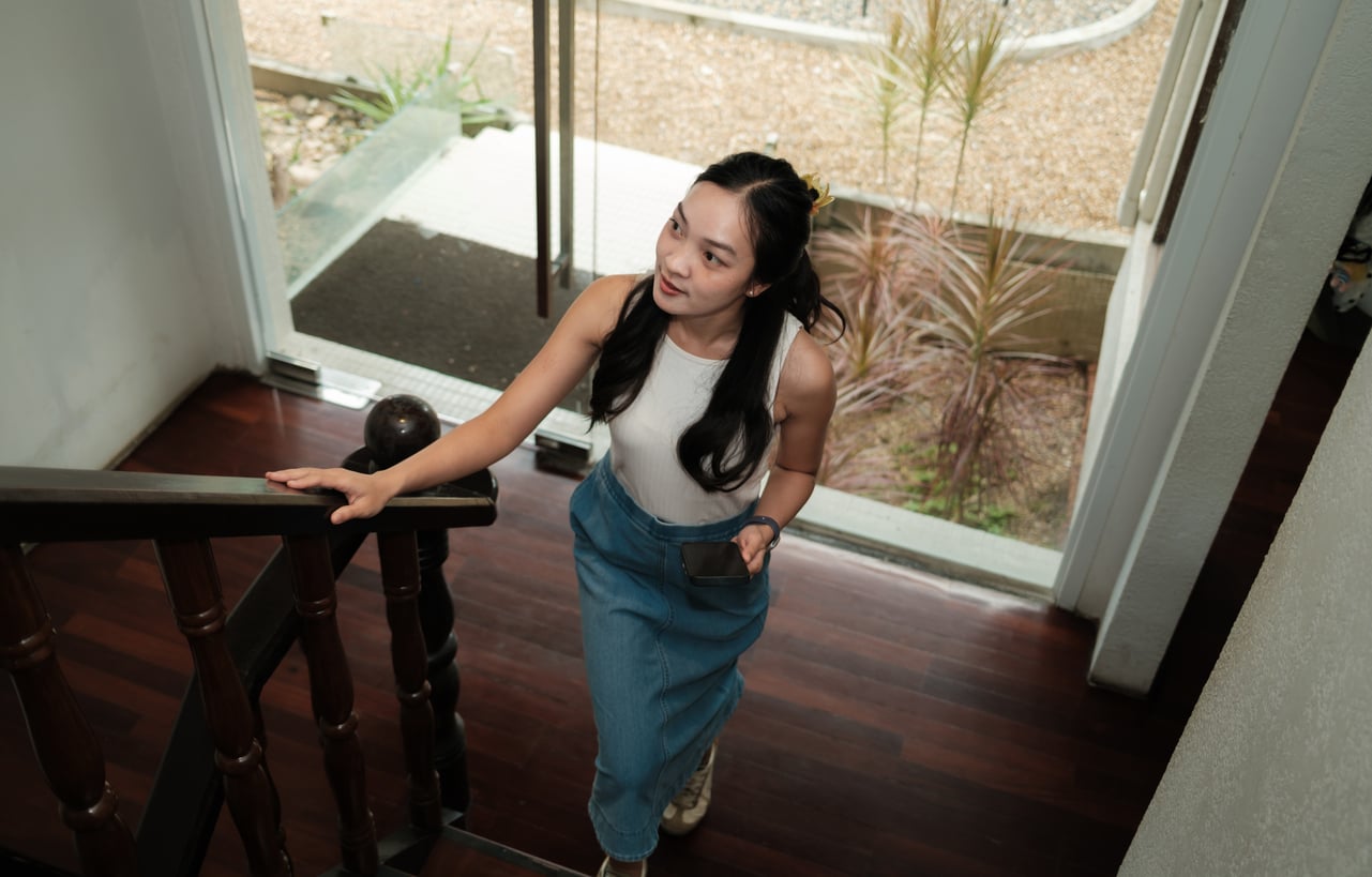 A woman with long dark hair, wearing a white sleeveless top and blue pants, holds a smartphone while walking up a wooden staircase in a modern home filled with natural light—a moment that stands in stark contrast to life before smartphones.