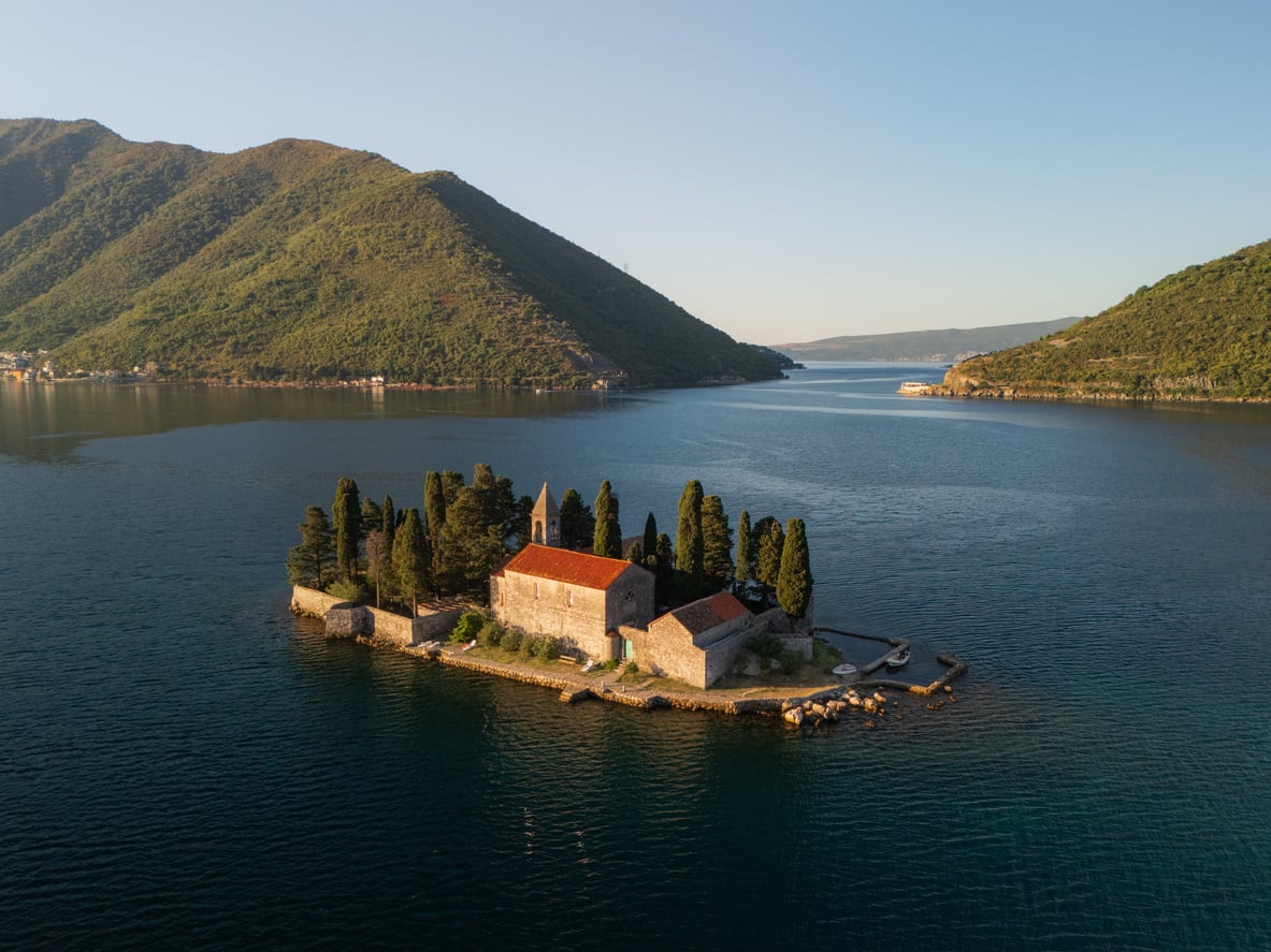 A small island with a stone building and tall trees, reminiscent of charming beach towns, surrounded by blue water and green hills under a clear sky.
