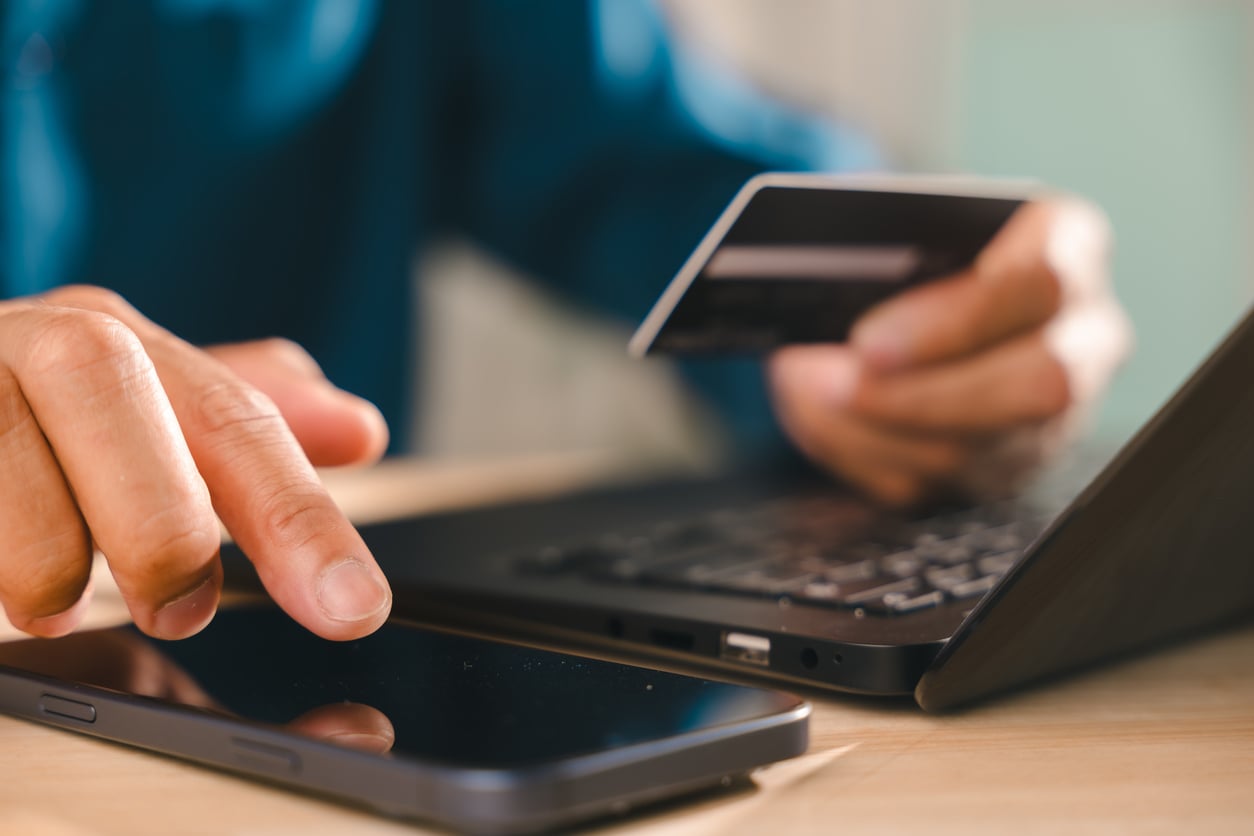 A person uses a smartphone and laptop while holding a credit card, suggesting an online purchase or digital payment.