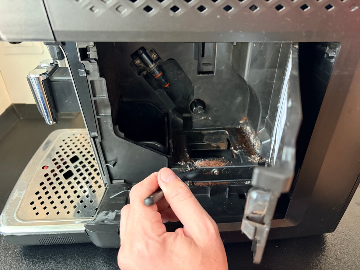A hand holds a cleaning brush inside the open compartment of a coffee machine, removing coffee grounds and residue from the interior components—a step that can help avoid issues common in products that don't last.