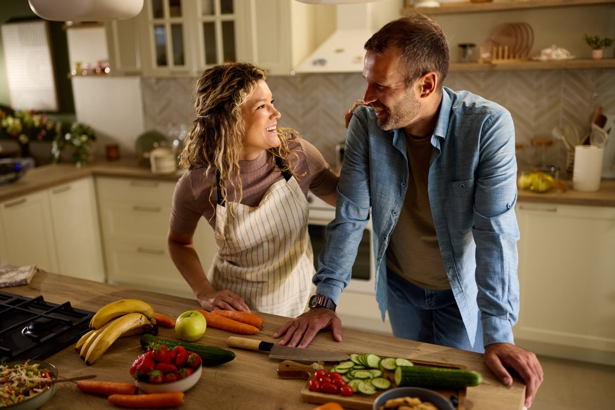 A smiling couple stands in a kitchen together. The woman wears an apron, and they are surrounded by fresh vegetables and fruit on the countertop, appearing to enjoy each other's company while preparing food.