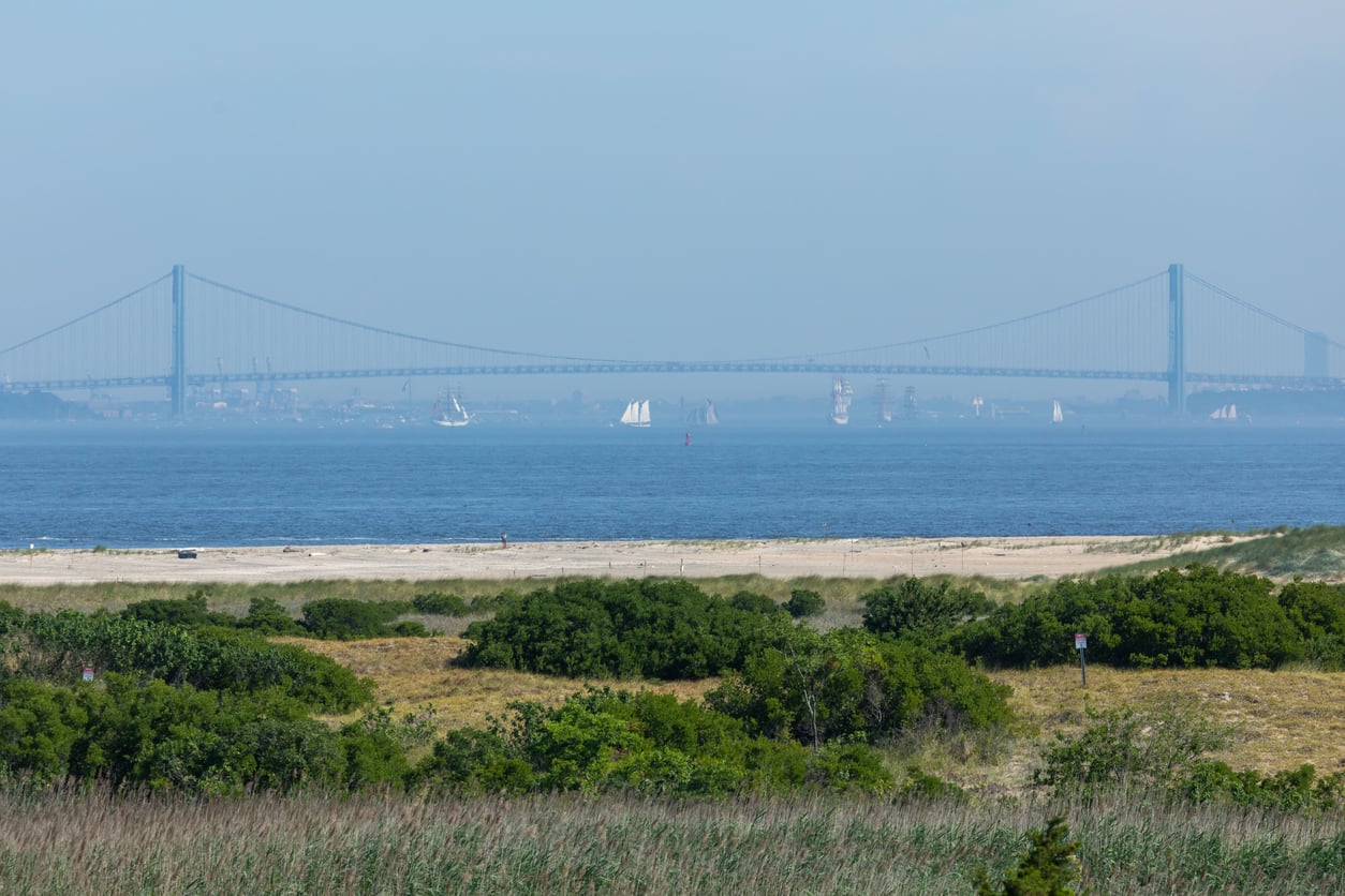 A wide view of an East Coast beach with green shrubs, blue water, and a distant suspension bridge. Several sailboats are visible on the water beneath a hazy sky.