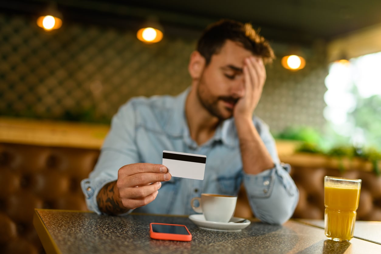 A man sits at a table with coffee, juice, and a phone, holding a credit card and covering his face with his hand, appearing stressed or worried—perhaps reflecting on the need for better money saving habits.