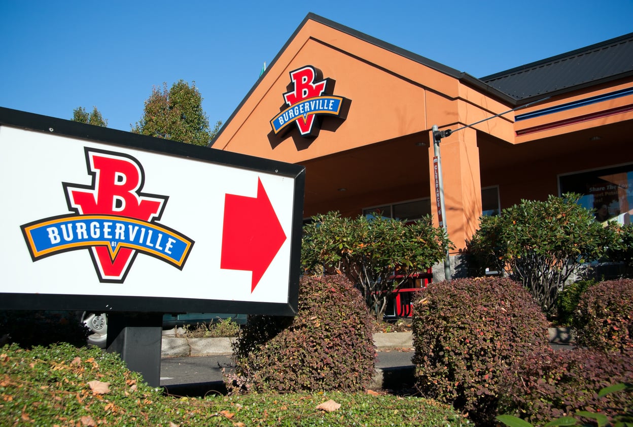 A Burgerville restaurant, part of the regional fast-food chains, stands with a large sign featuring the Burgerville logo and a red arrow pointing right, surrounded by bushes under a clear blue sky.