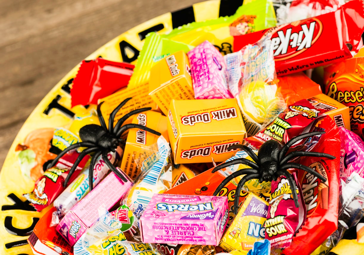 A colorful bowl of assorted Halloween candies, including Nerds, Milk Duds, Reese's, Kit Kat, Starburst, and Laffy Taffy, with two plastic black spiders on top.