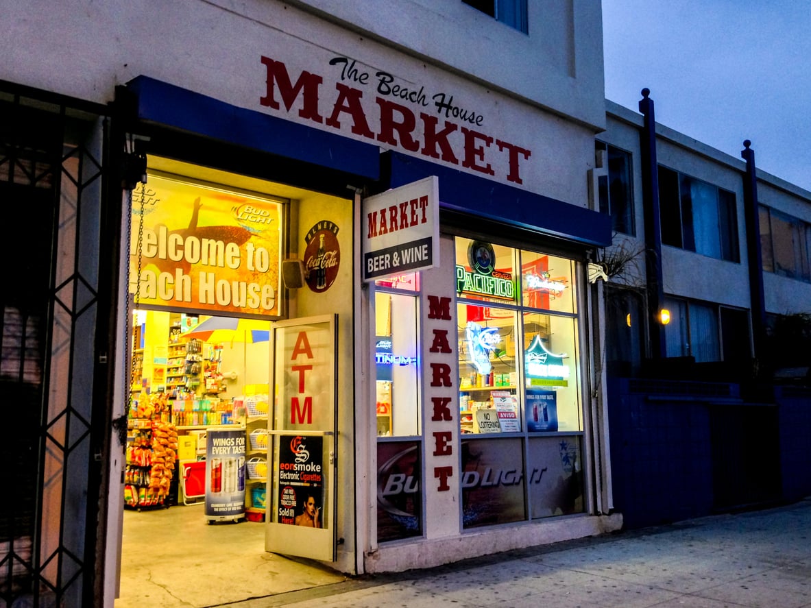 Exterior view of "The Beach House Market" at dusk with bright lights inside, an ATM sign, and assorted convenience store items visible—perfect for beach vacation savings. A "Welcome to Beach House" sign hangs inside.