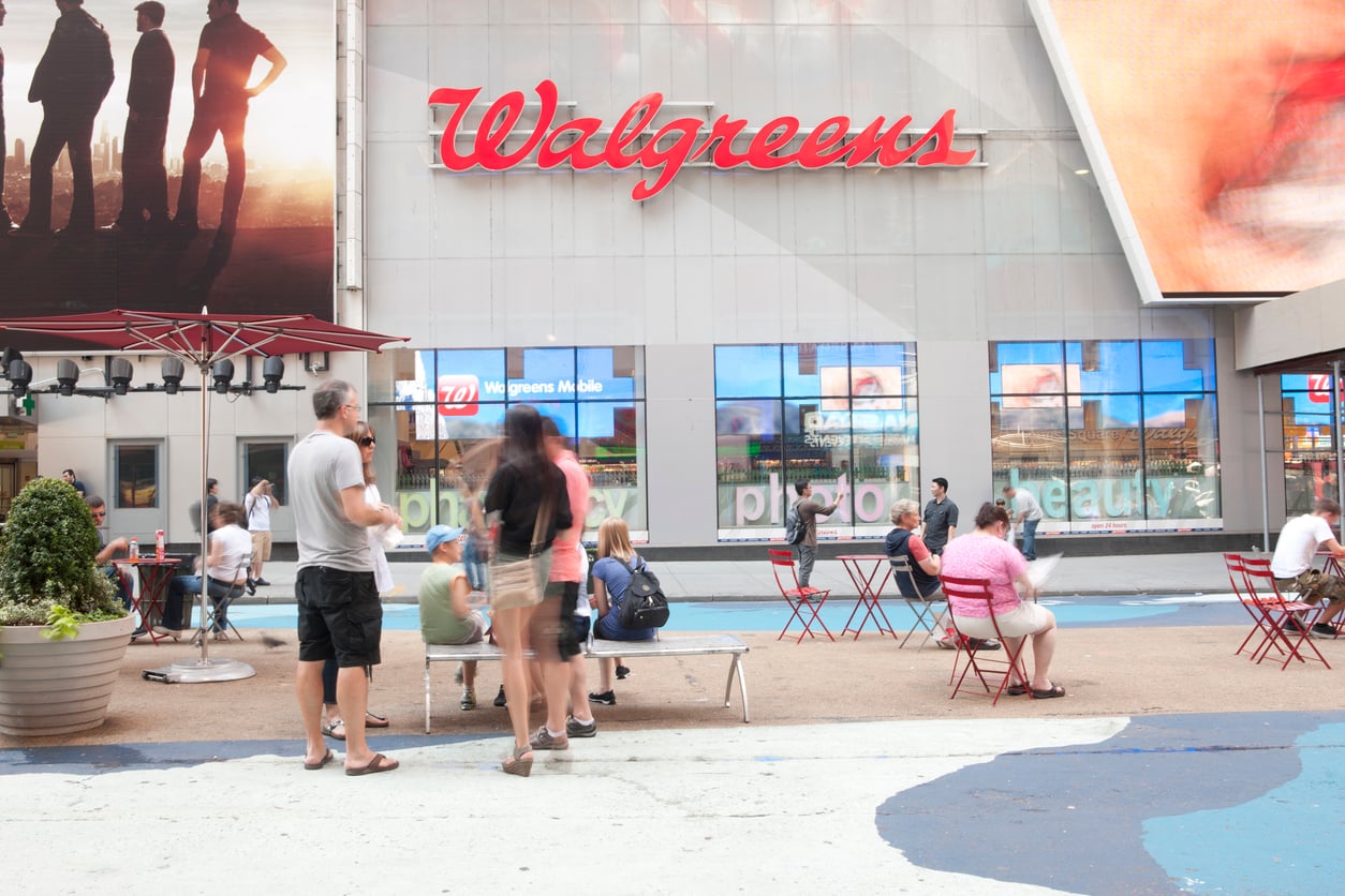 People sit and stand in an outdoor plaza in front of a Walgreens store. Red chairs and tables are scattered around, and large ads and signs are visible on the building.