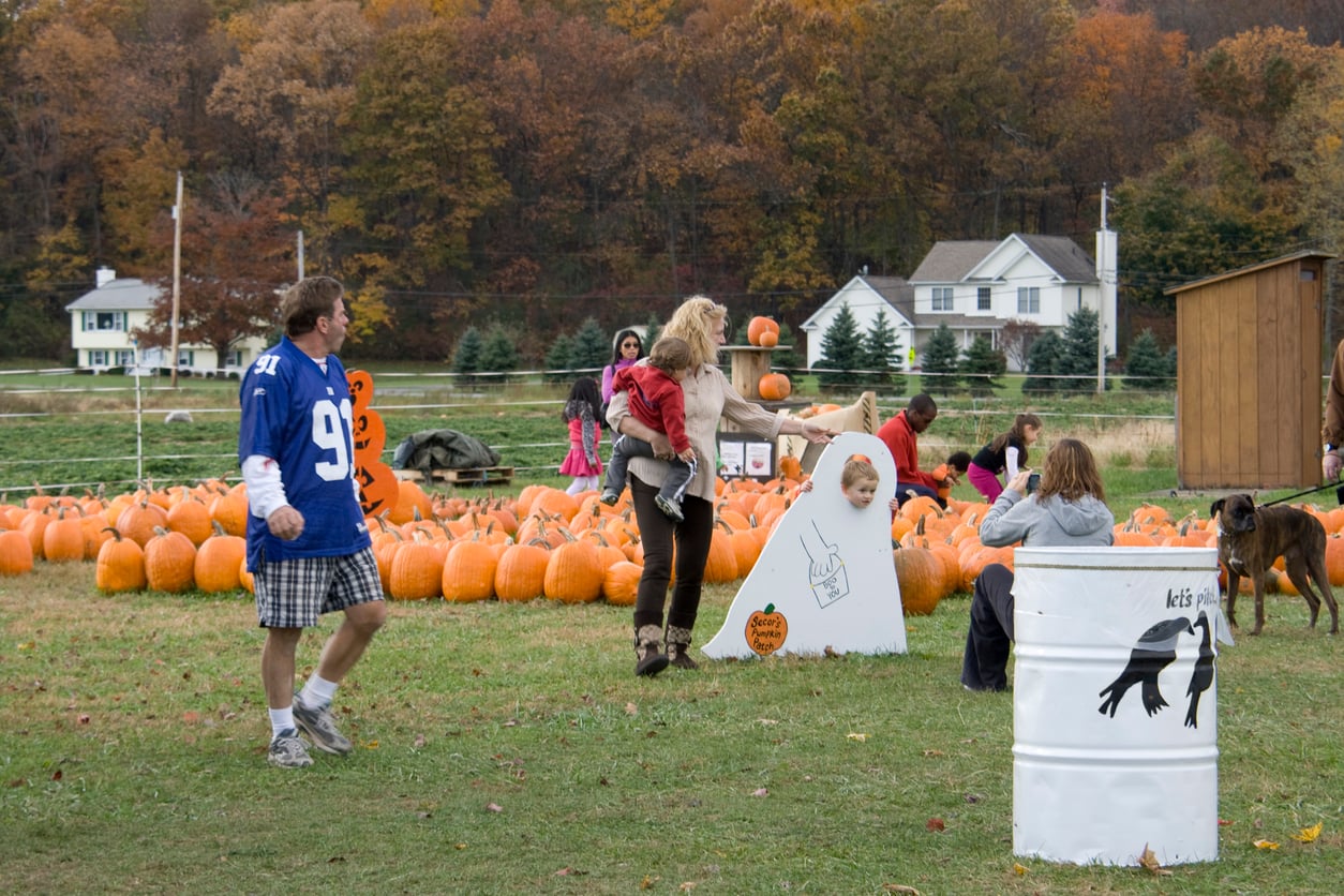 People enjoy a pumpkin patch filled with festive Halloween activities; a man in a blue jersey mingles nearby, kids pose with a ghost cutout, and a child rides on a woman’s back. Pumpkins are scattered around, while trees adorned with vibrant fall foliage create the perfect seasonal backdrop.