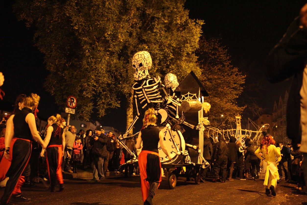 A nighttime parade in one of the best trick or treat towns in America features large skeleton puppets, performers in red and black costumes, and a crowd watching under trees and streetlights.