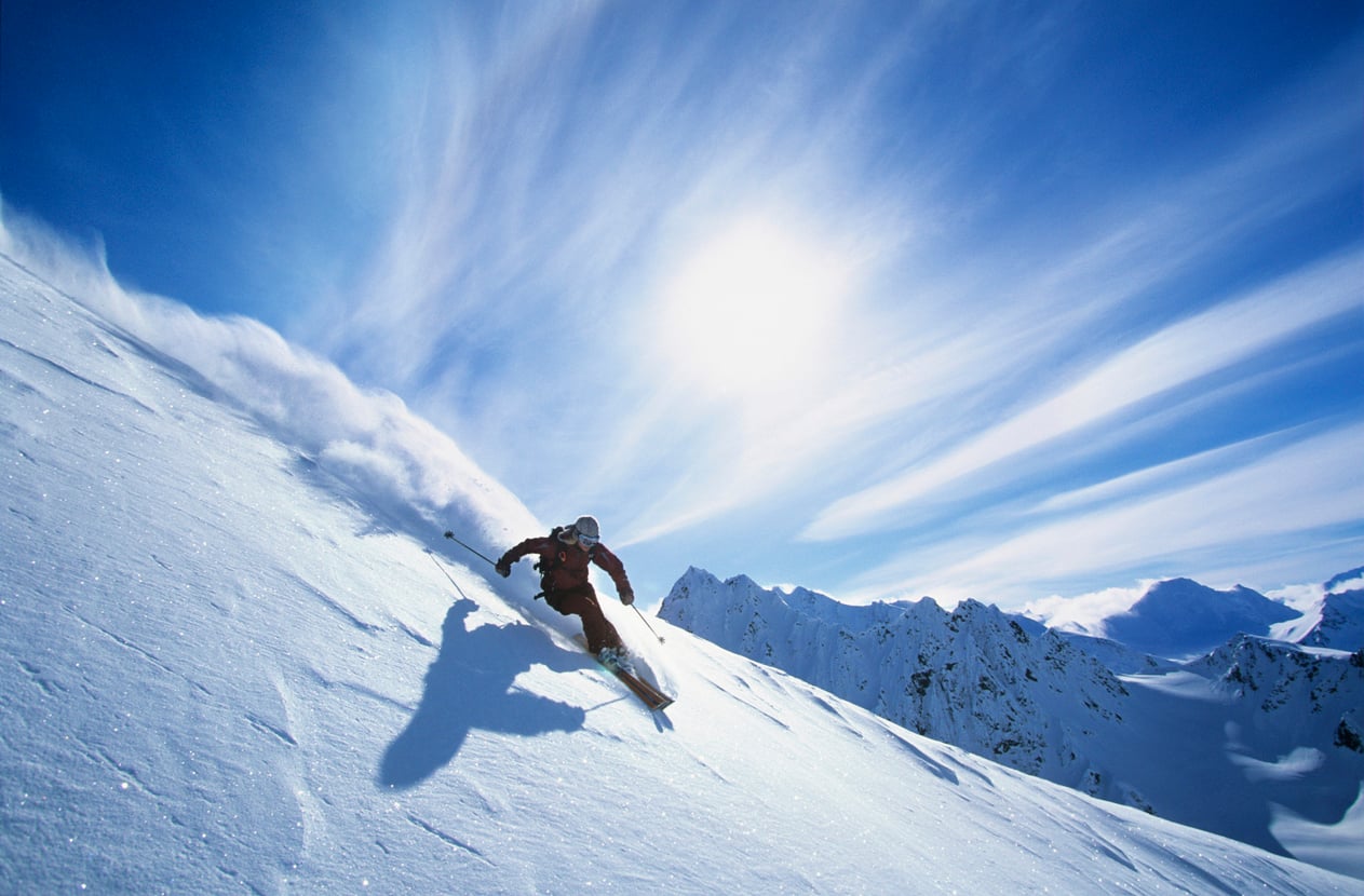 A skier in dark gear descends a steep, snowy mountain slope under a bright sun and blue sky with streaked clouds, leaving a trail of powder behind—a thrilling example of one of the world’s most expensive hobbies. Jagged peaks are visible in the background.