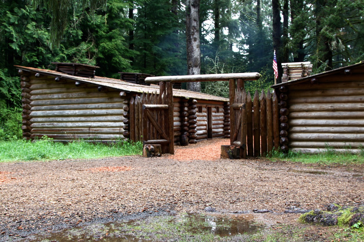 A rustic wooden log fort with a gated entrance stands among tall trees, with an American flag on the right. The ground is wet and muddy, and the area is surrounded by dense green forest—an evocative scene that captures the allure of unique travel destinations on the West Coast.