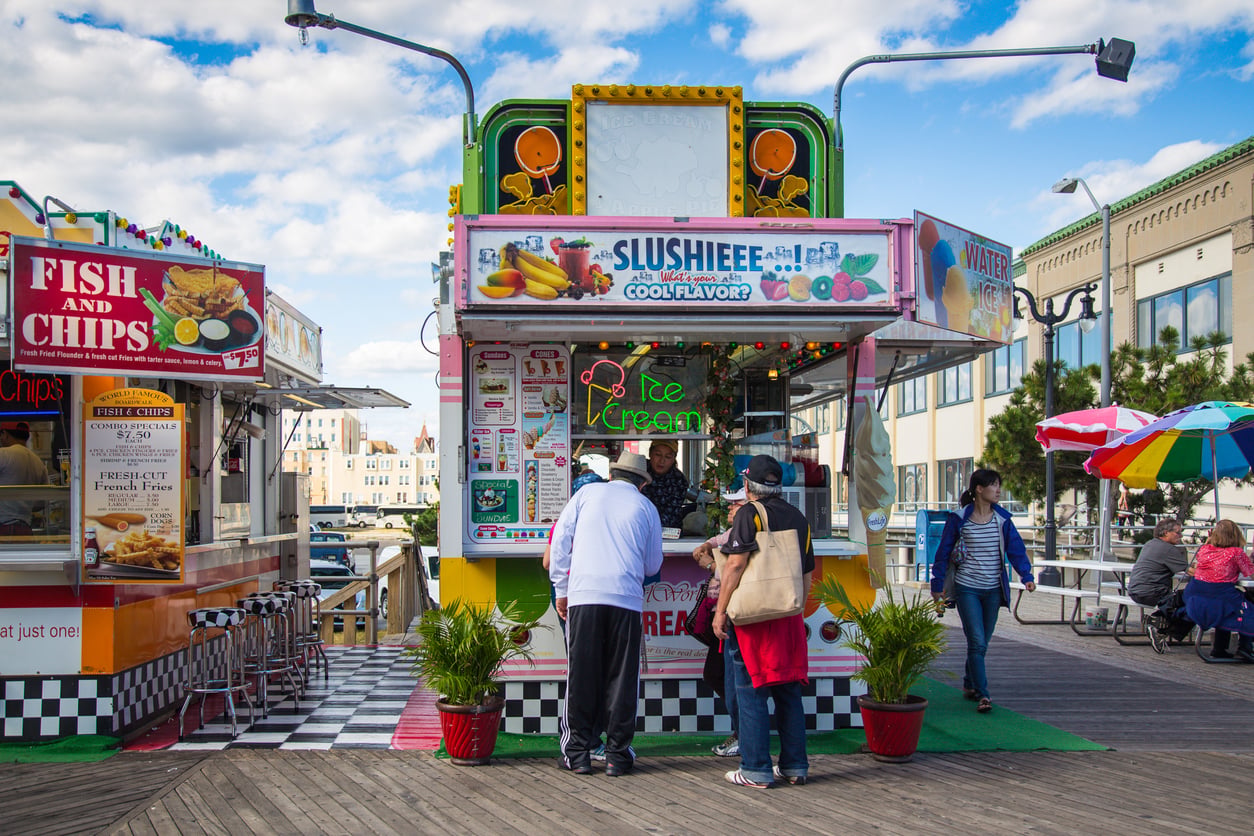 A colorful ice cream and slushie stand on a boardwalk serves customers, with a fish and chips stall next to it. People enjoy beach vacation savings as they stroll by or relax at tables under umbrellas beneath a partly cloudy sky.