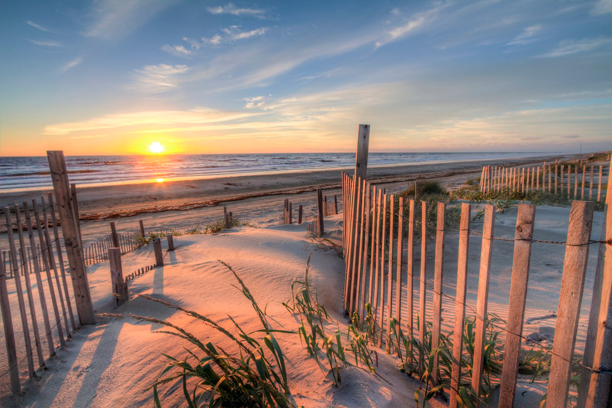 Wooden fences line sandy dunes with some beach grass, leading to a wide sandy beach at sunrise or sunset. The sun is low on the horizon over a calm ocean under a sky with wispy clouds.