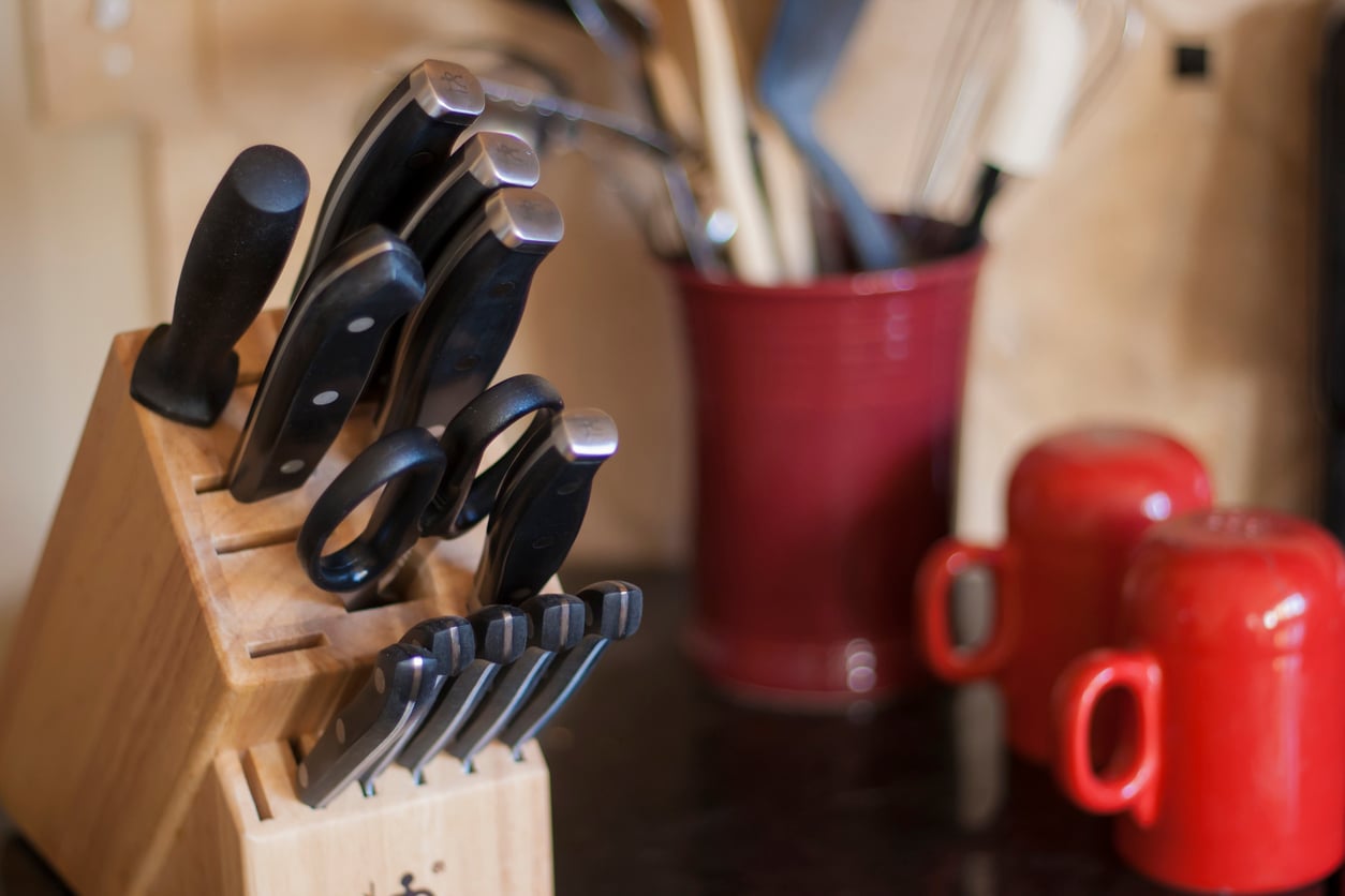 A wooden knife block with knives and scissors sits on a kitchen counter, surrounded by essential kitchen items. In the background are a red utensil holder with kitchen tools and two red mugs.