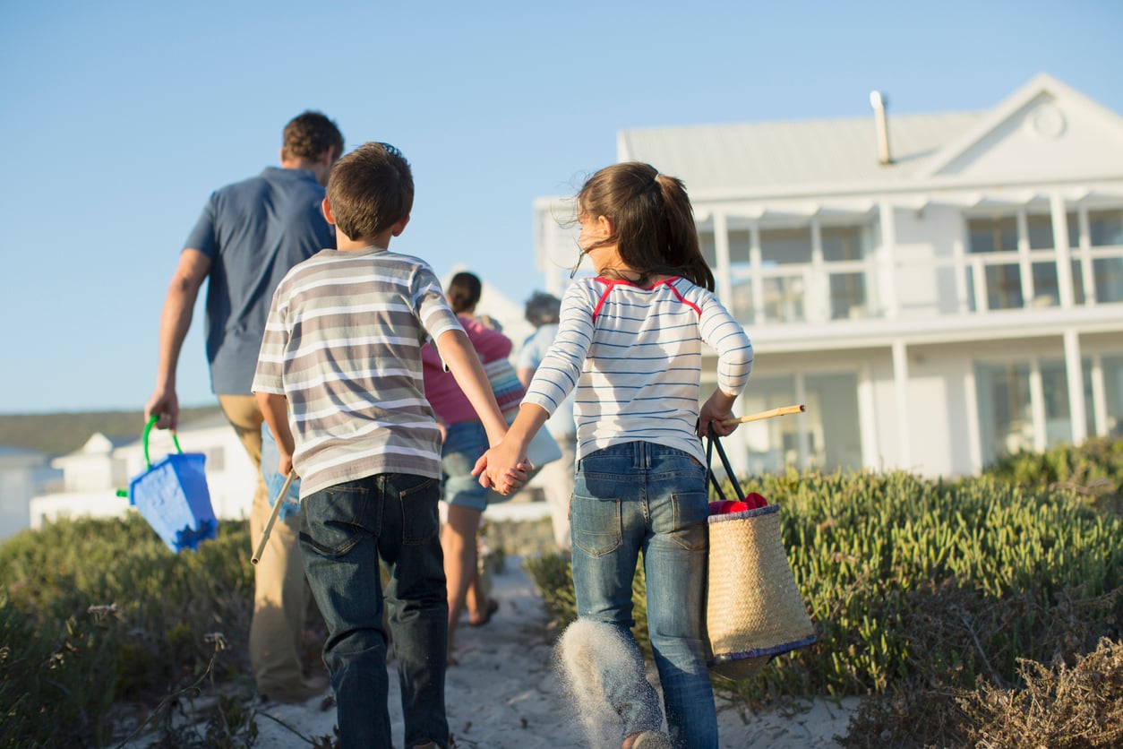 Two children holding hands and carrying beach bags walk with their family toward a white beach house on a sunny day, enjoying the excitement of vacation swaps. The group is walking on a sandy path surrounded by greenery.