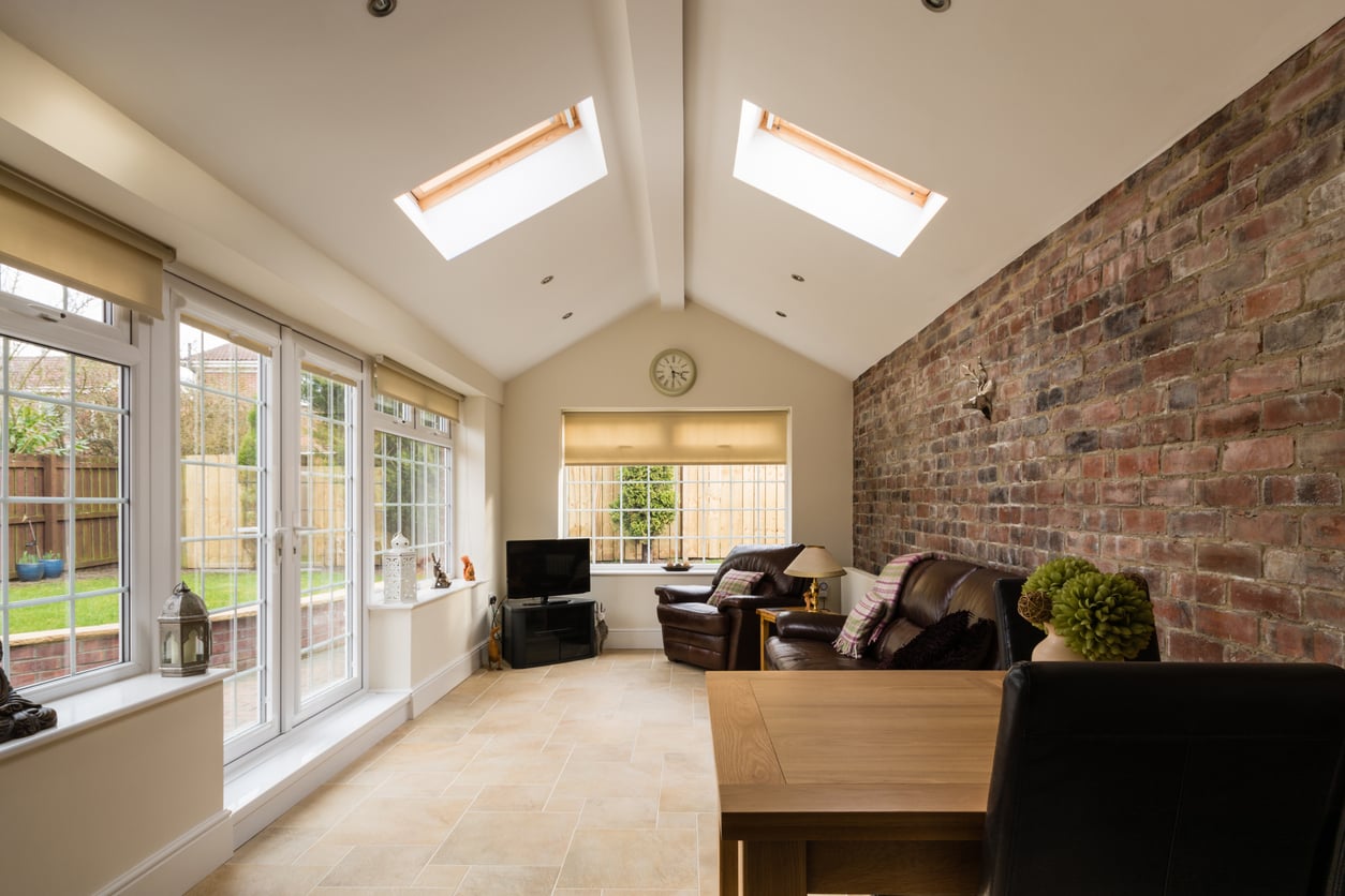 A bright sunroom with two skylights, large windows, a brick accent wall, leather sofas, a TV, a clock, and a wooden desk with a chair. The room overlooks a fenced backyard garden.